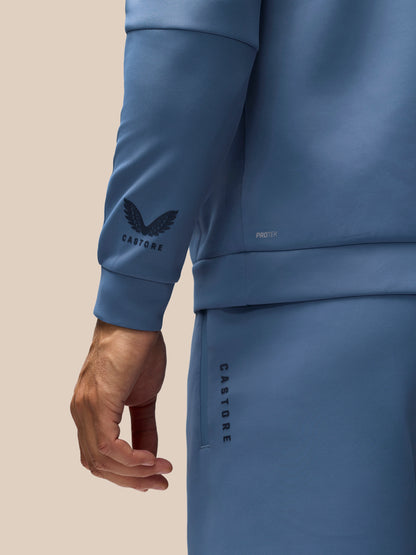 Close-up of a person wearing a dusty blue Castore tracksuit, showing the sleeve with a black winged logo and the word 'CASTORE', the side pocket with vertical 'CASTORE' text, and part of the hand against a plain beige background.