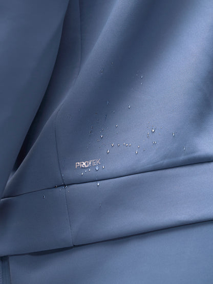 Close-up of a blue-gray fabric surface with water droplets beading on it and a small visible PROTEK logo near the lower left area.