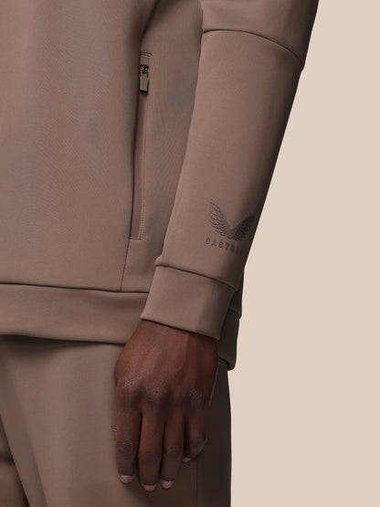 Close-up of a person wearing a beige tracksuit with a brand logo on the sleeve against a beige background