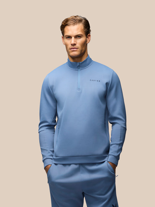 A man modeling a light blue athletic tracksuit, featuring a quarter-zip long-sleeve top and matching pants, against a solid beige background.