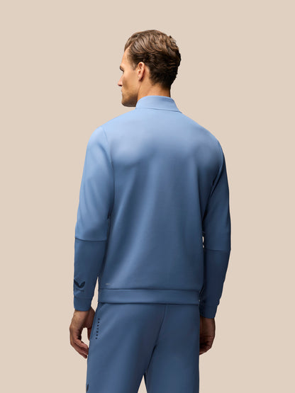 A man shown from the back wearing a matching dusty blue tracksuit, featuring a mock neck long-sleeve top and matching pants, against a solid beige background.