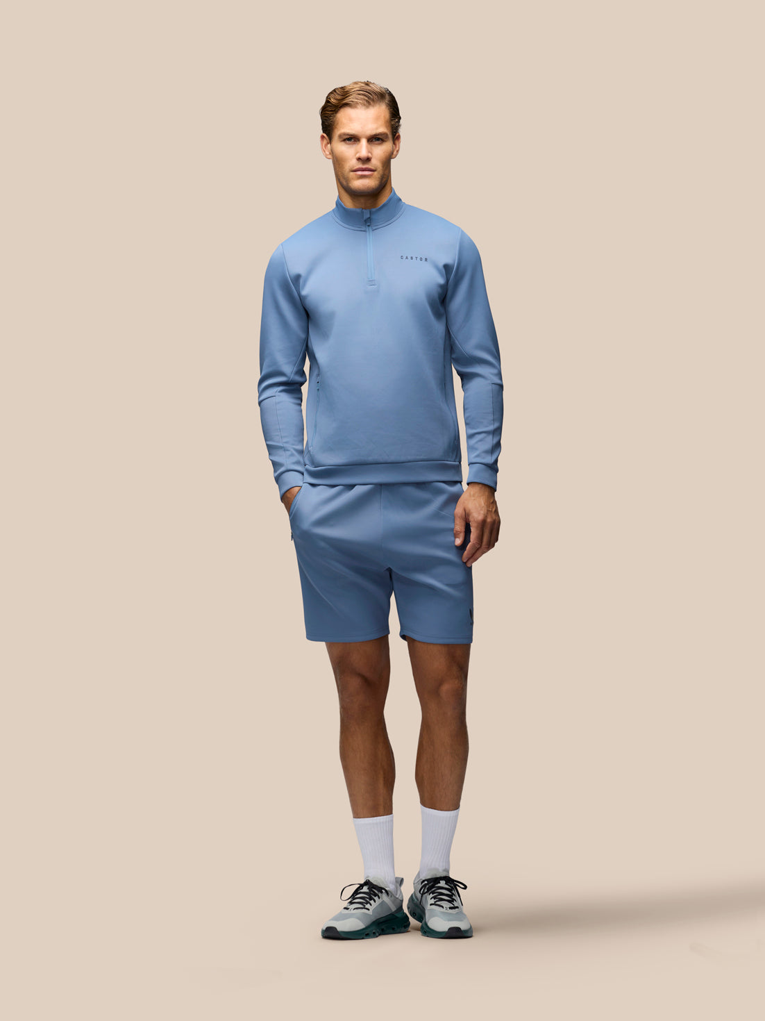 A male model stands against a solid beige studio background wearing a light blue athletic half-zip pullover and matching shorts, paired with white socks and dark athletic shoes.