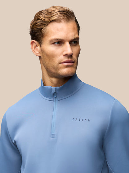 A man wearing a light blue quarter-zip pullover sweatshirt with the brand name 'CASTORE' printed on the chest, set against a solid beige background.