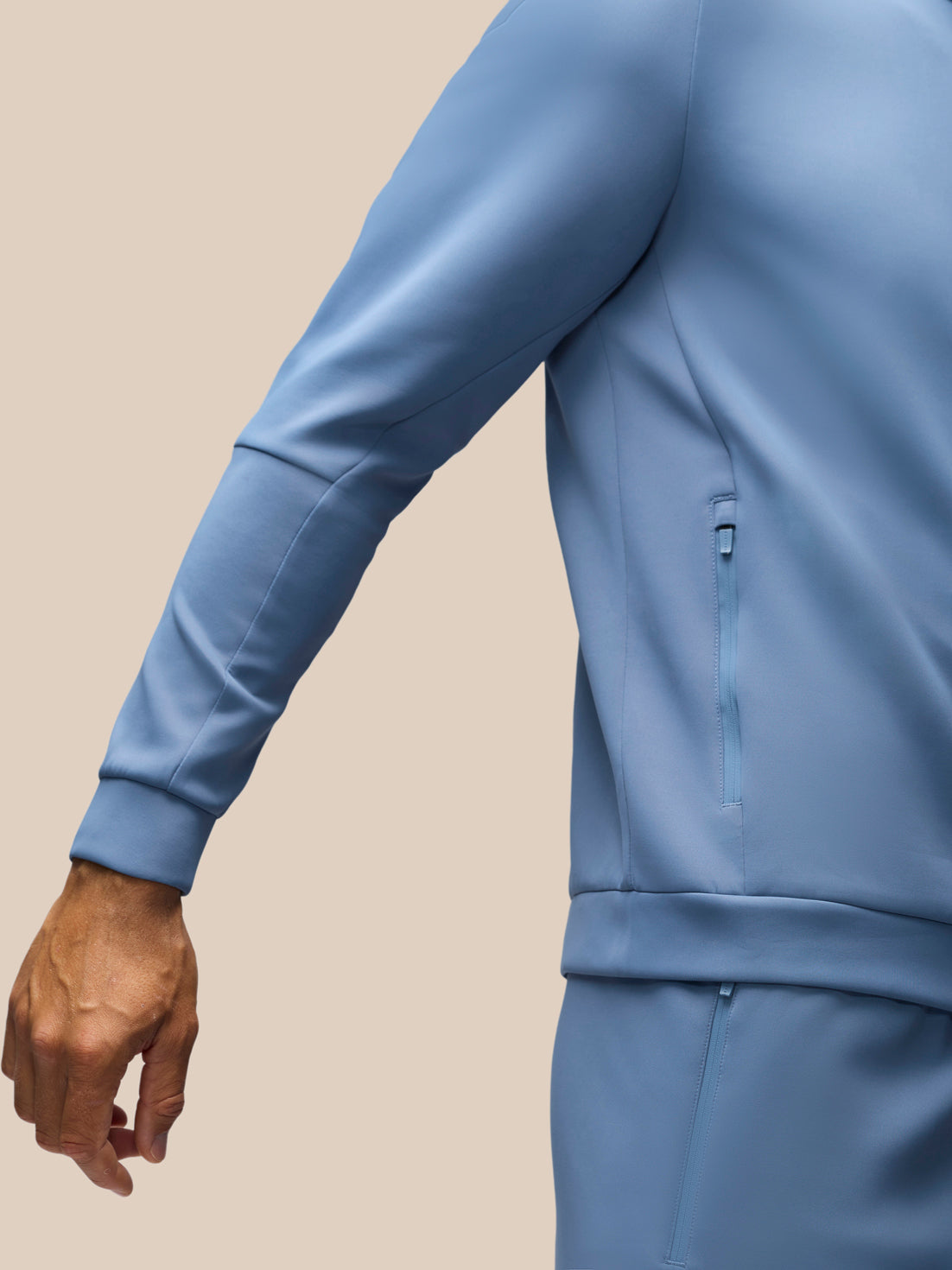 A close-up of a person wearing a light blue long-sleeve zip-front jacket and matching bottoms against a plain beige studio background.