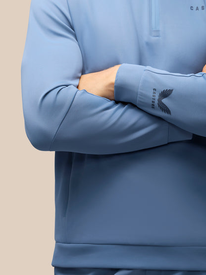 A close-up of a person wearing a light blue long-sleeve quarter-zip pullover with a dark gray wing logo and the brand name 'CASTORE' printed on the left sleeve cuff.