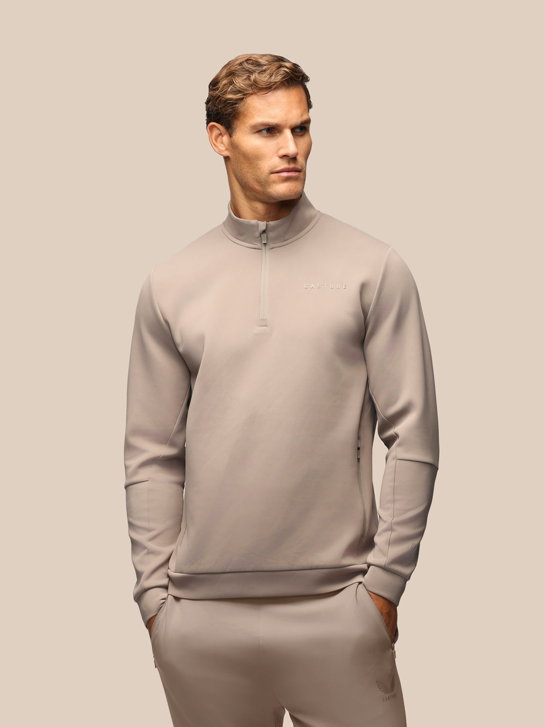 A man wearing a light taupe-gray quarter-zip pullover sweatshirt and matching sweatpants, standing against a solid beige background.