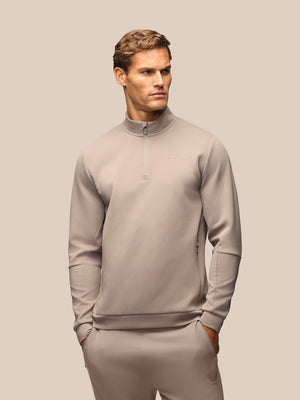 A man wearing a light taupe-gray quarter-zip pullover sweatshirt and matching sweatpants, standing against a solid beige background.