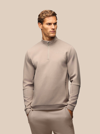 A man wearing a light taupe-gray quarter-zip pullover sweatshirt and matching sweatpants, standing against a solid beige background.