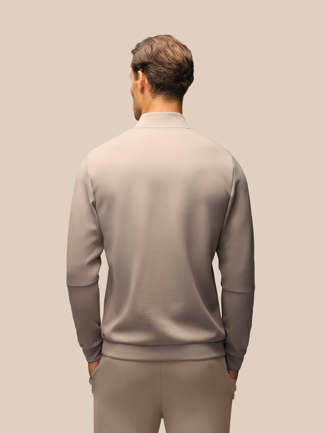 A man viewed from behind wearing a light taupe-beige mock neck long-sleeve top and matching trousers against a solid beige background.