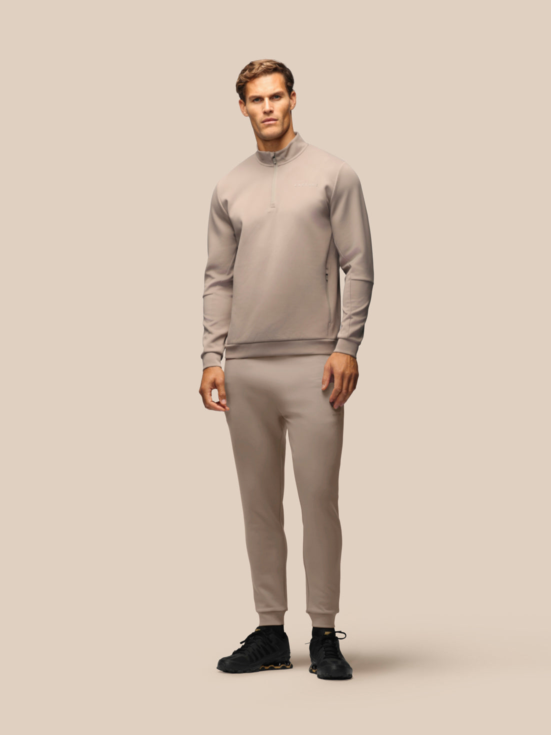 A male model wearing a matching light taupe-gray athletic tracksuit, consisting of a long-sleeve quarter-zip pullover and matching jogger pants, paired with black sneakers, against a solid beige studio background.