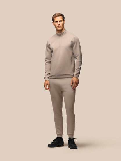 A male model wearing a matching light taupe-gray athletic tracksuit, consisting of a long-sleeve quarter-zip pullover and matching jogger pants, paired with black sneakers, against a solid beige studio background.
