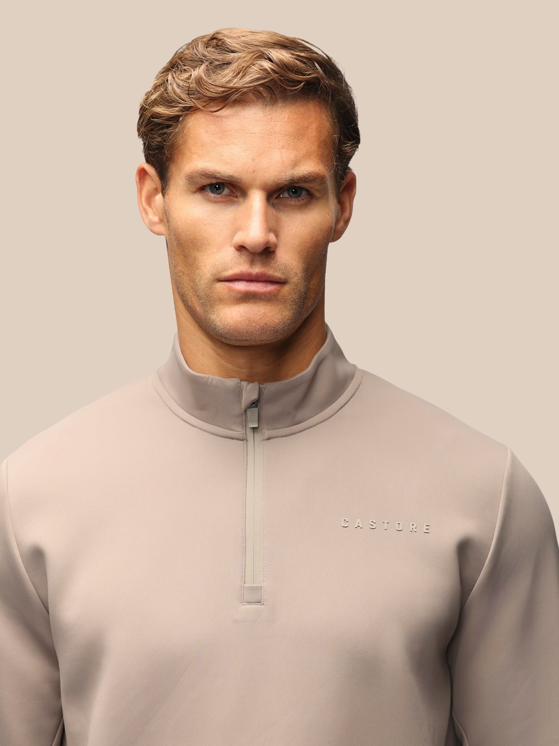 A man wearing a light taupe quarter-zip pullover sweatshirt with the brand name 'CASTORE' subtly printed on the left chest, set against a solid beige background.