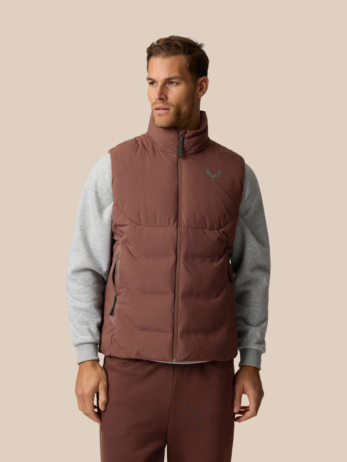 A man wearing a brown puffer vest over a gray long-sleeve shirt and matching brown pants, standing against a solid beige background.