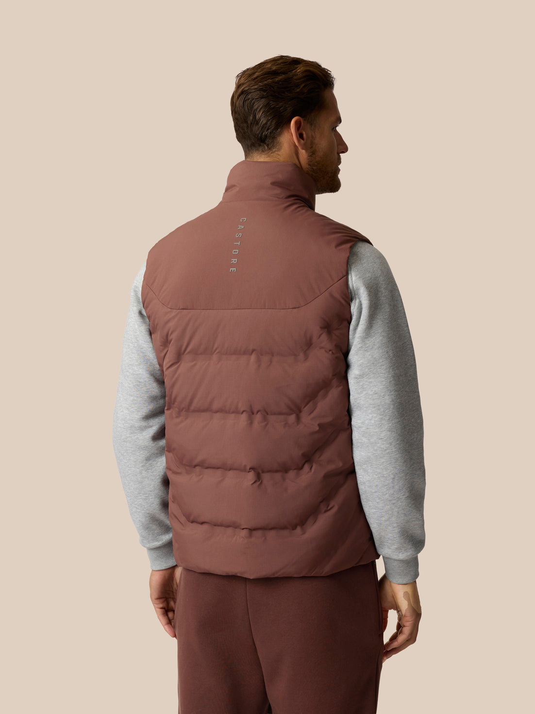 A man shown from the back wearing a brown puffer vest over a gray long-sleeve shirt and brown pants, against a solid beige background.