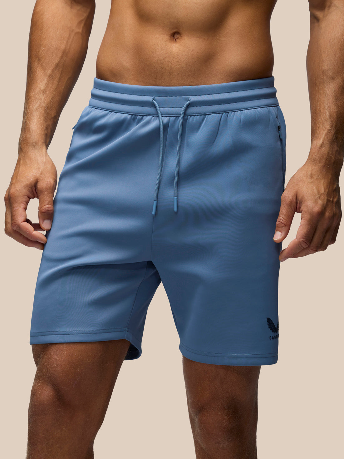 A close-up of a man wearing blue athletic shorts with a drawstring waist and a small black logo near the lower right leg, against a plain beige background.