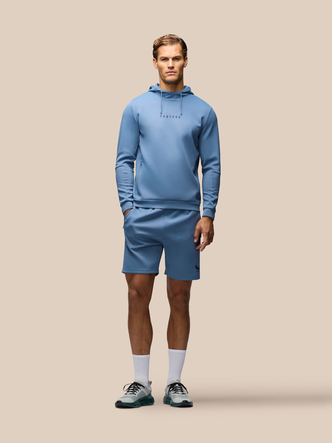 A male model stands against a solid beige background wearing a matching light blue athletic hoodie and shorts set, white crew socks, and gray running shoes.