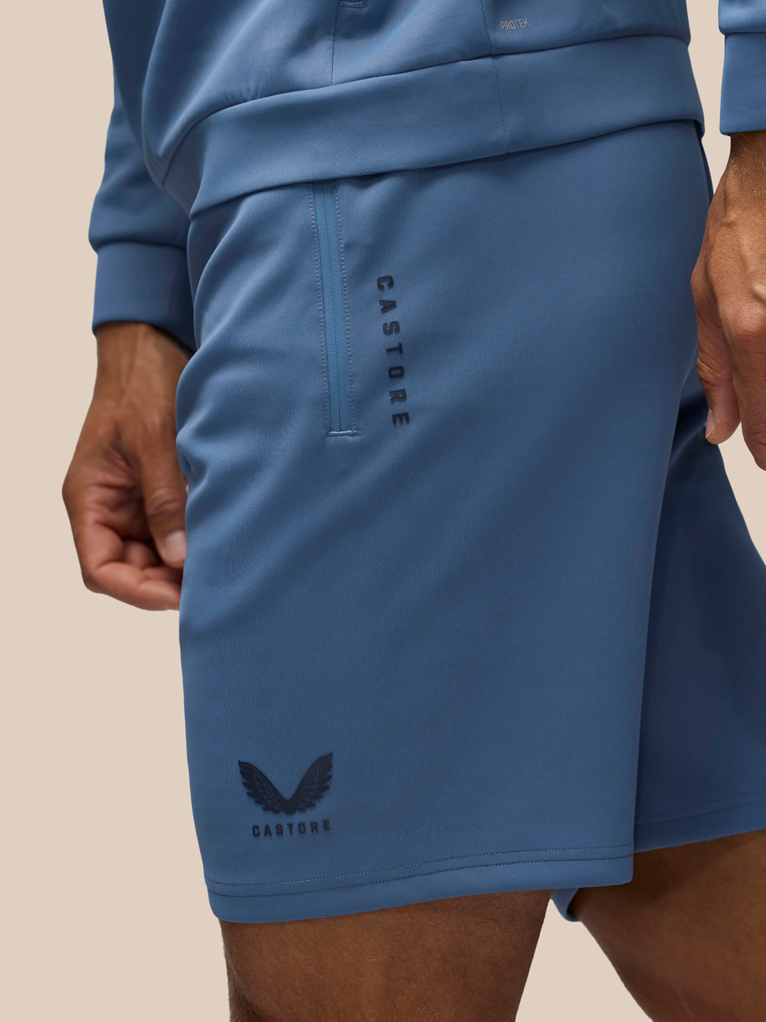 Close-up of a person wearing blue athletic shorts with a vertical zip pocket and black Castore branding, paired with a matching blue top against a plain beige background.