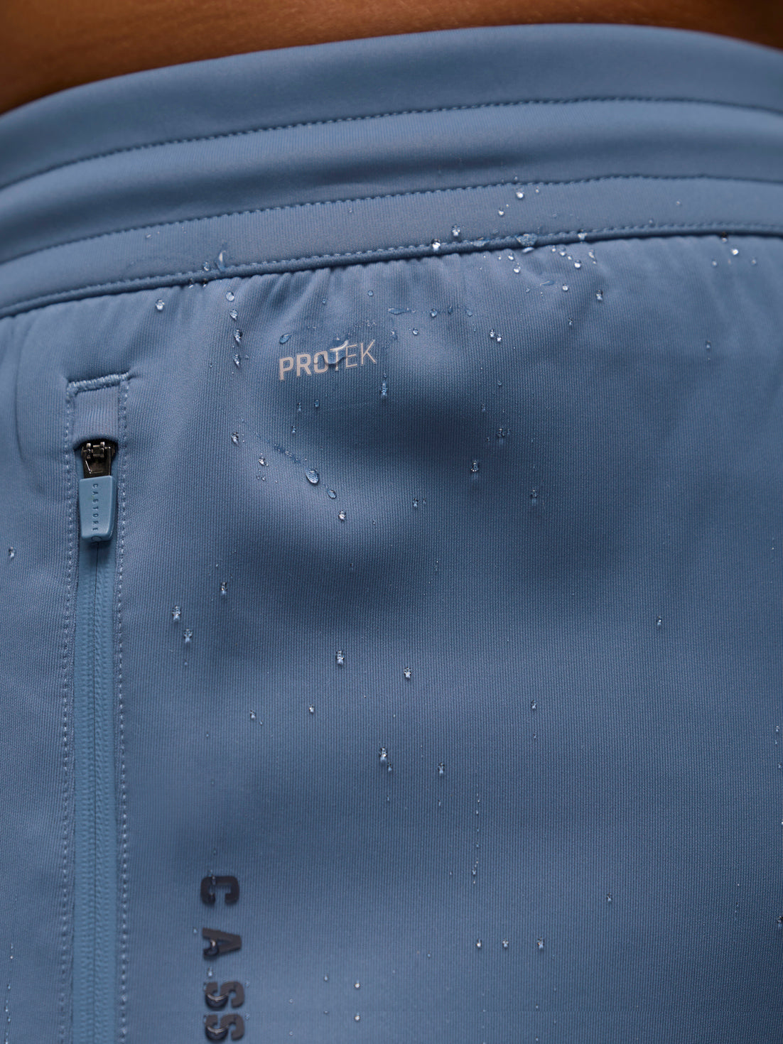Close-up of a light blue athletic garment showing the PROTEK water-repellant finish with water droplets on the surface and a vertical zipper pocket.