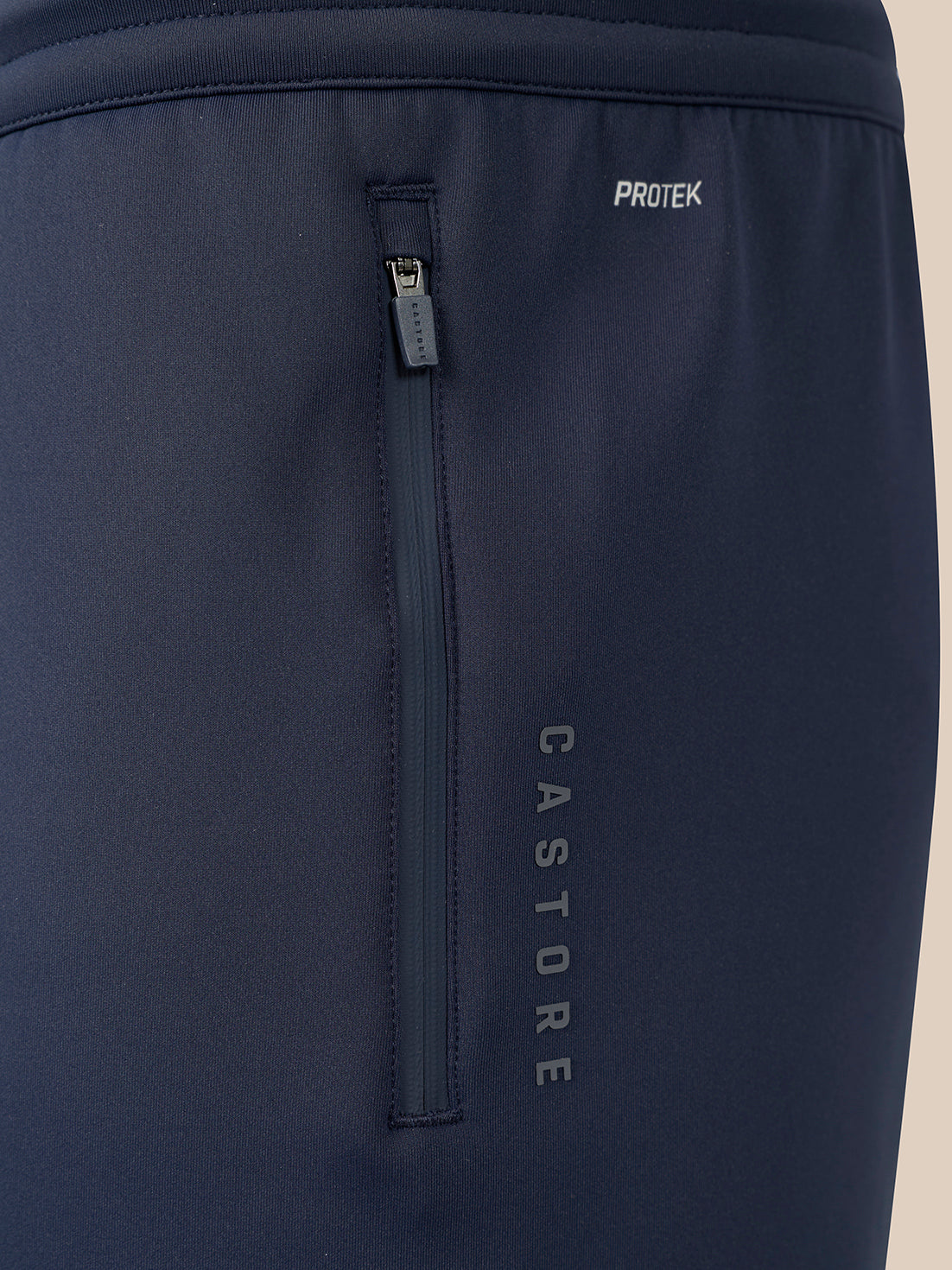 Close-up of dark navy athletic pants showing a vertical zippered pocket with the vertical brand name CASTORE and a small horizontal zip pocket with the brand name PROTEK.