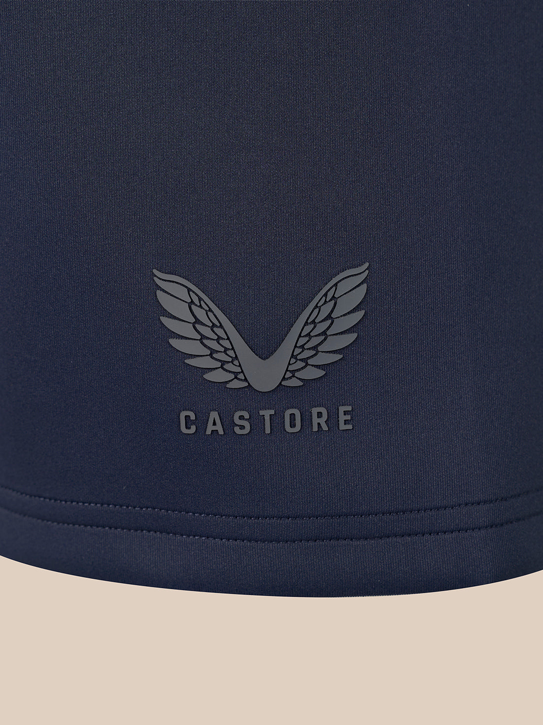 Close-up of a navy blue garment with a tonal gray winged logo and the word CASTORE centered on the chest, against a light beige background.