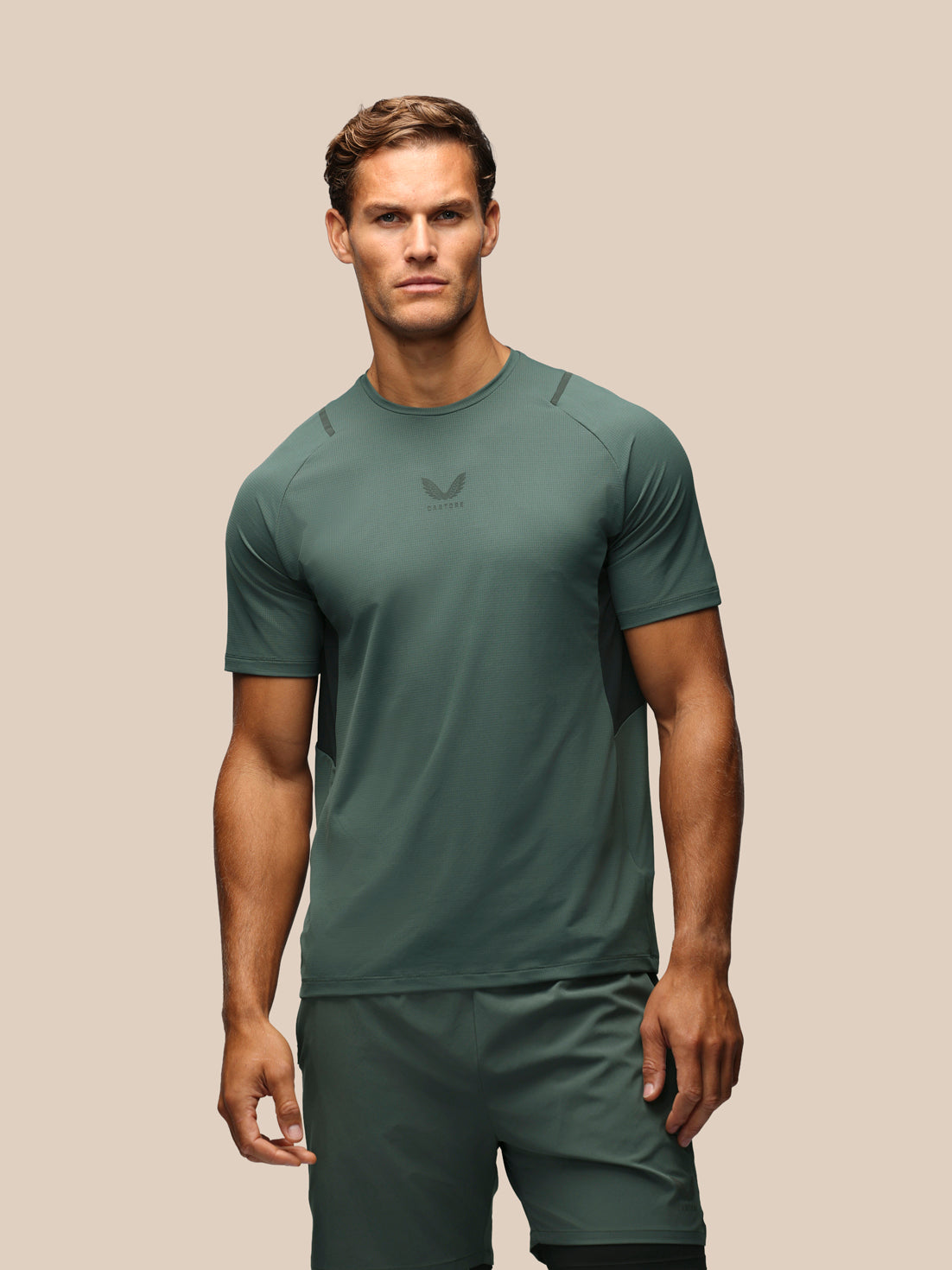 A man modeling a dark green athletic short-sleeve shirt and matching shorts against a solid beige background.