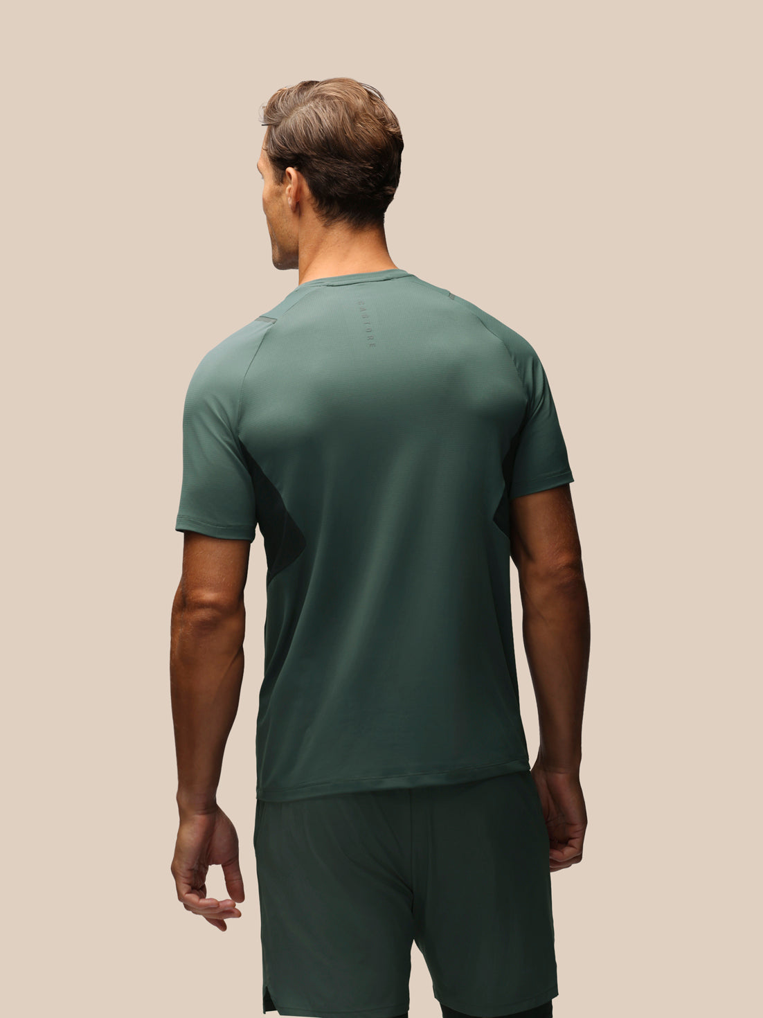 A man viewed from behind wearing a dark green athletic t-shirt and matching shorts against a solid beige background.