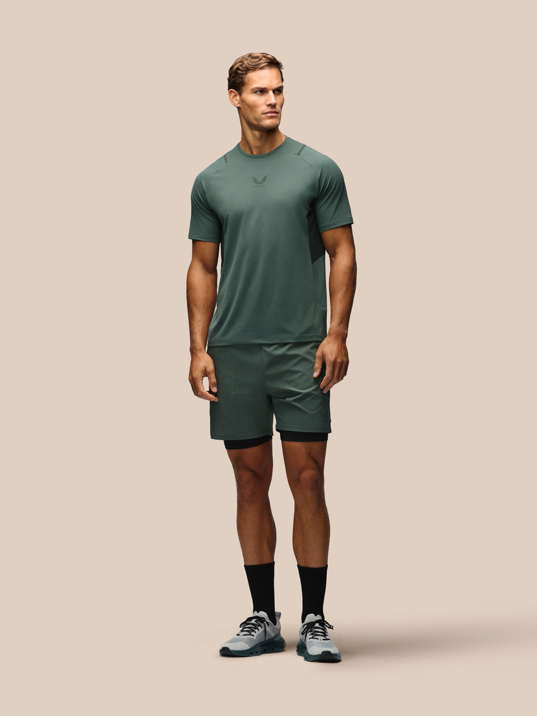 A man modeling a matching athletic set consisting of a short-sleeve crewneck t-shirt and matching shorts in a muted green color, paired with black socks and gray running shoes, against a solid beige background.