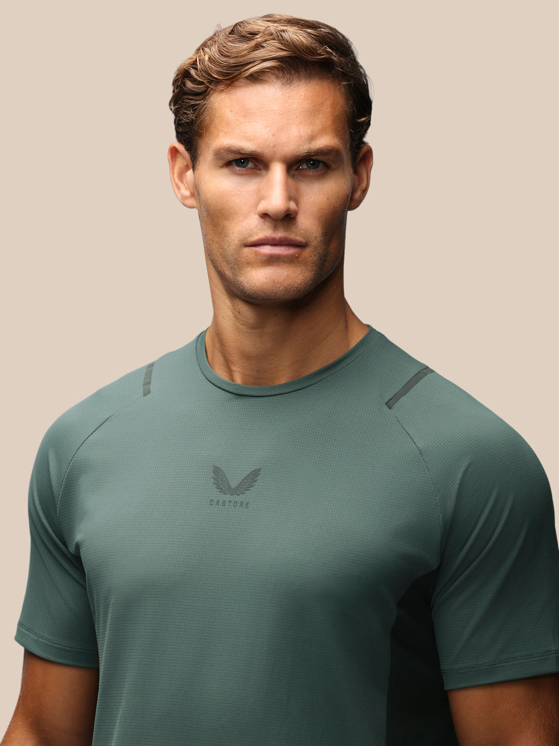 A man wearing a dark green athletic t-shirt with a subtle wing logo and the brand name 'CASTORE' printed on the chest, set against a solid beige background.