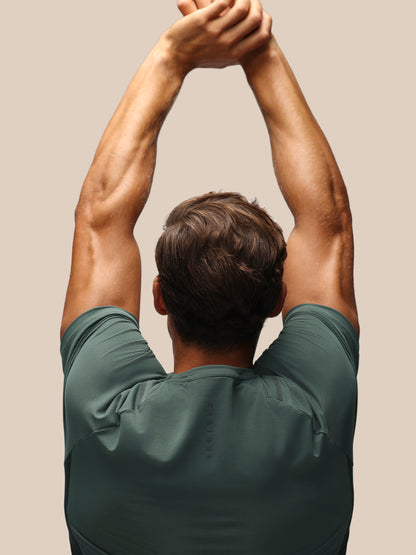 A man viewed from behind wearing a dark green athletic t-shirt, stretching his arms overhead against a solid beige background.