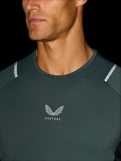 Close-up of a man wearing a dark green Castore athletic shirt with a textured waffle knit, featuring a silver wing logo and 'CASTORE' text on the chest, and reflective silver shoulder stripes.