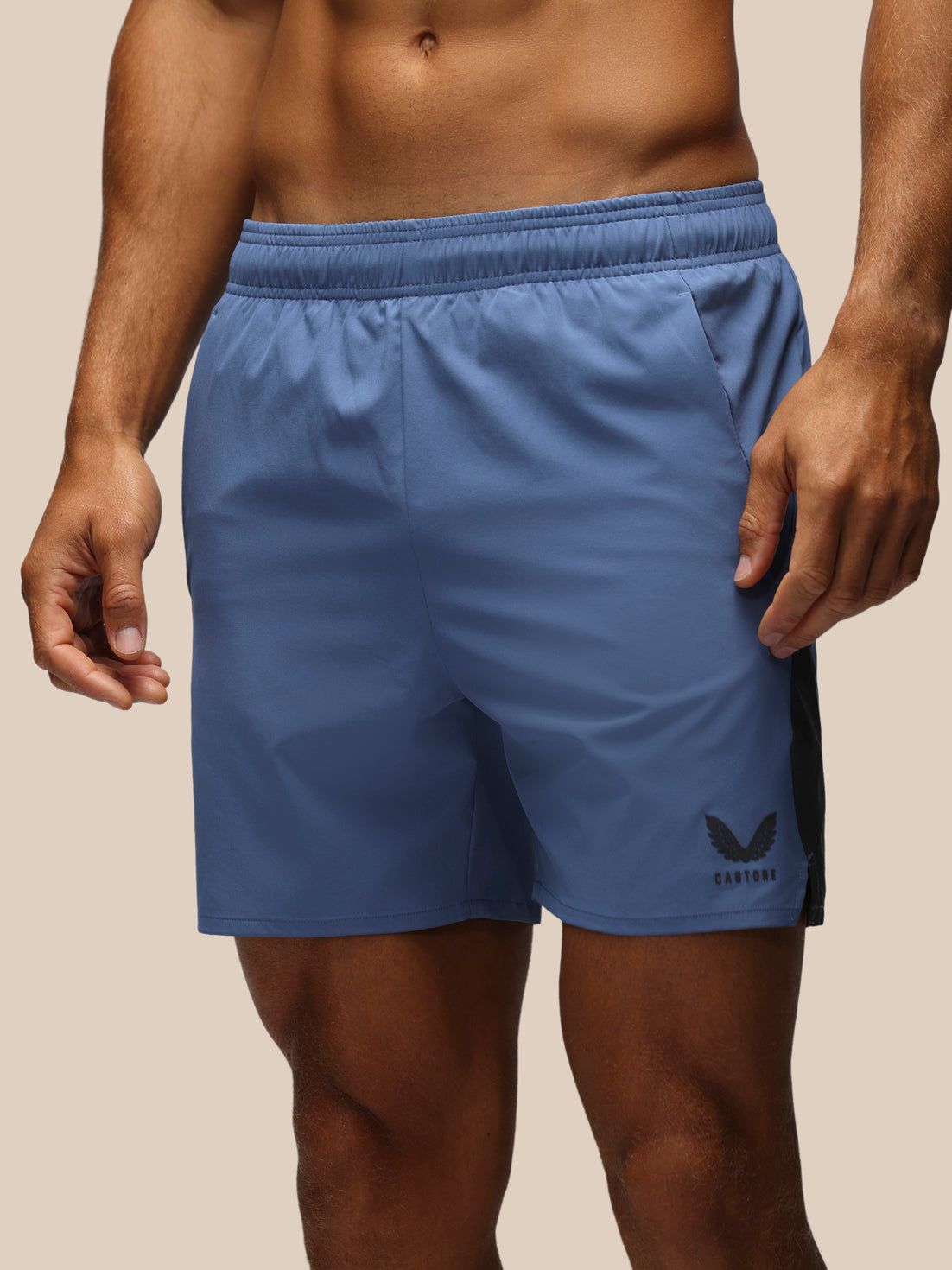 A close-up of a man wearing blue athletic shorts with a black side logo, shown from the chest to the upper thighs against a plain beige studio background.