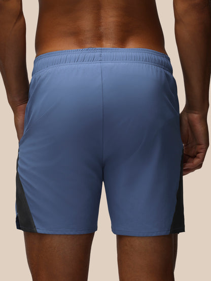 Rear view of a person wearing athletic shorts in a dusty medium blue color, with dark side panel accents, against a solid beige background.