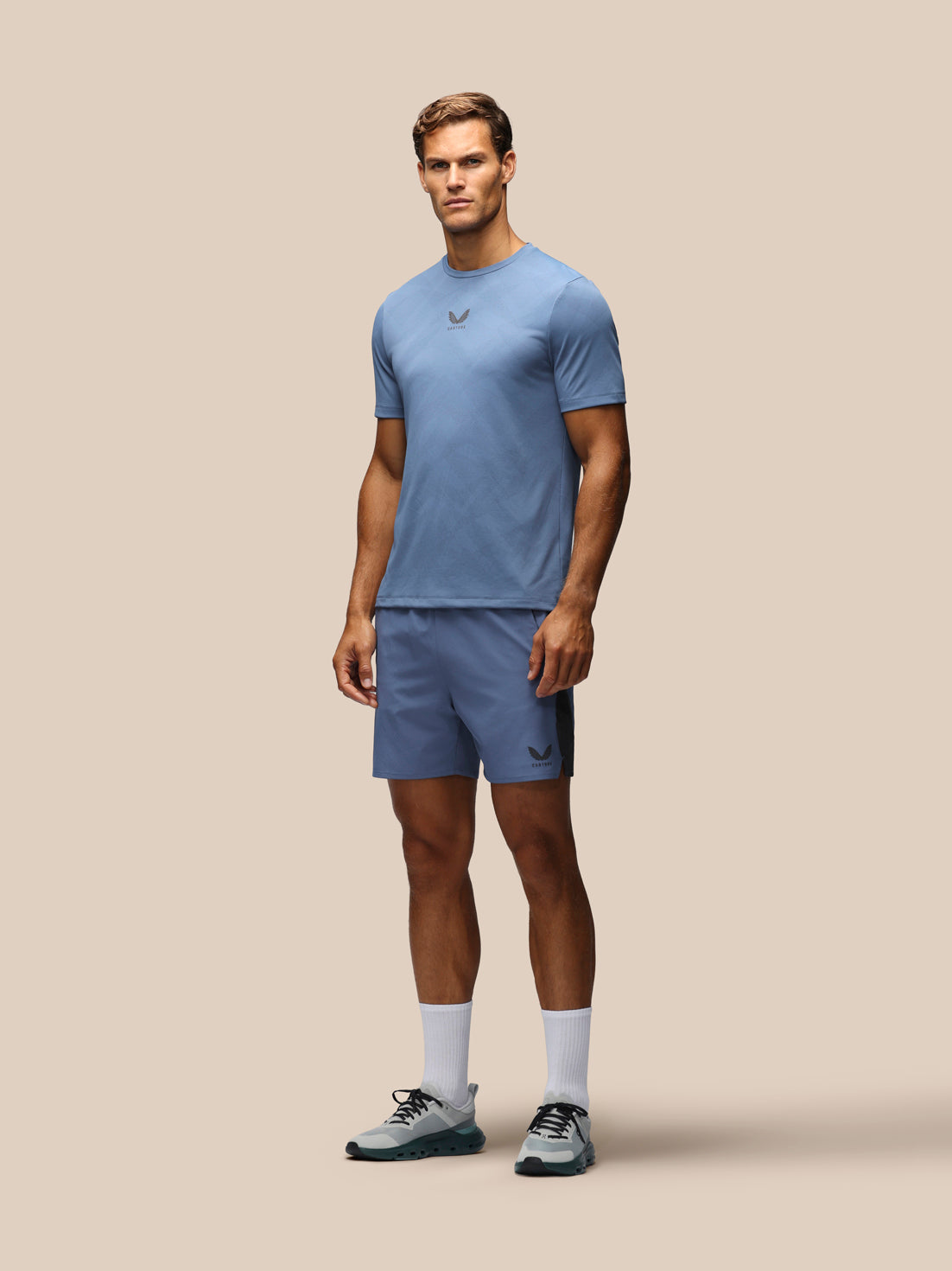 A male model stands against a solid beige background wearing a matching light blue athletic t-shirt and shorts set, white crew socks, and gray running shoes. The set features a small dark logo on the chest and left leg.