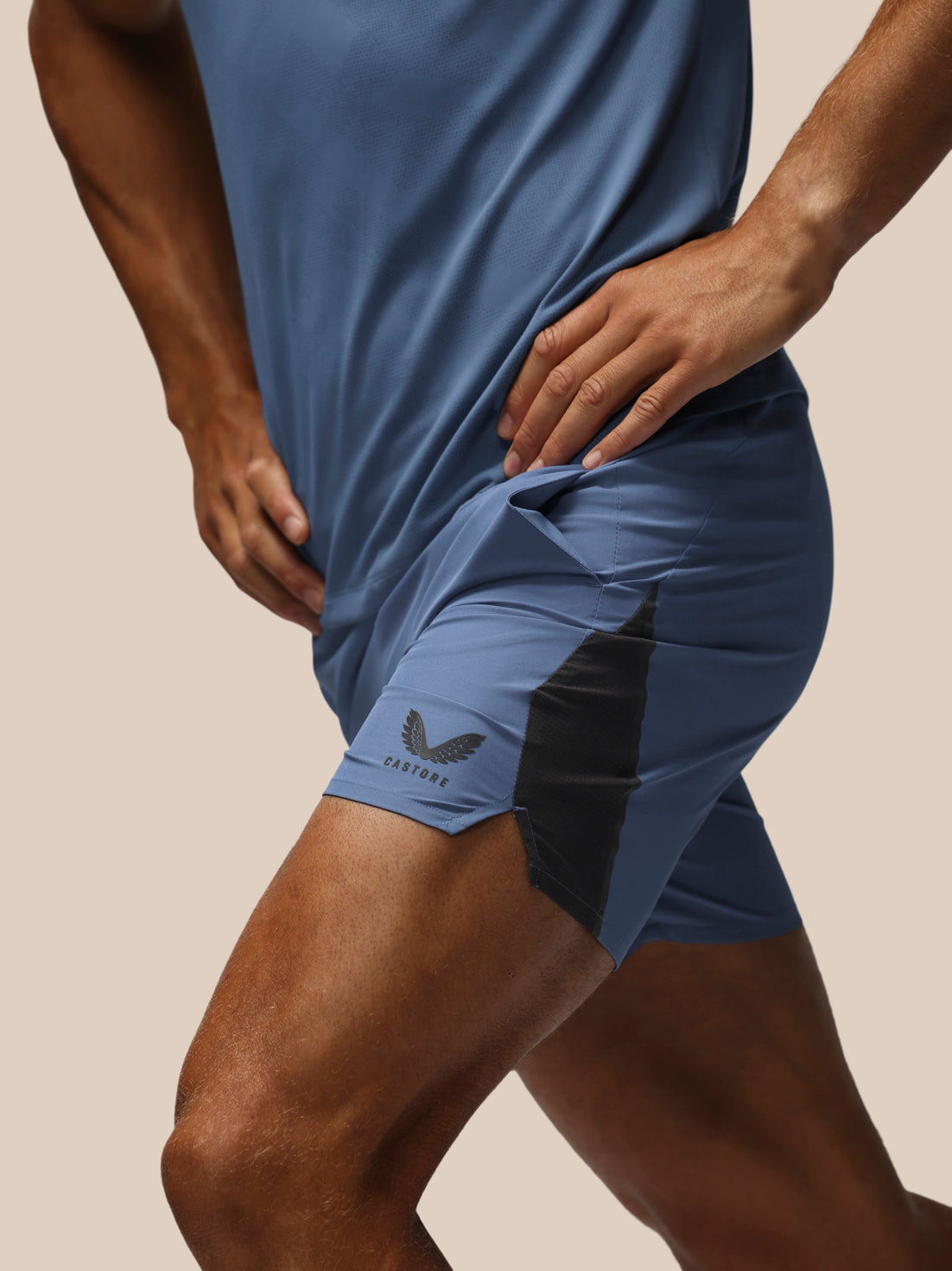 Close-up of a person wearing blue athletic shorts with a black side panel and a visible castore logo, paired with a matching blue top against a plain beige studio background.