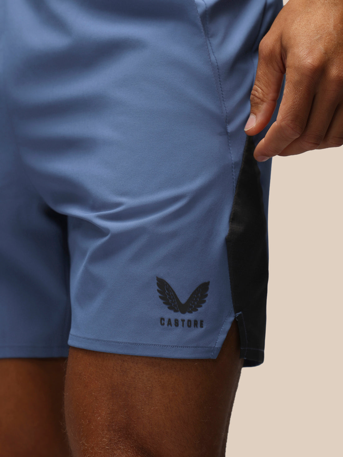Close-up of a person wearing blue athletic shorts with a black side panel and a black wing logo with the word CASTORE near the hem, against a plain light beige background.