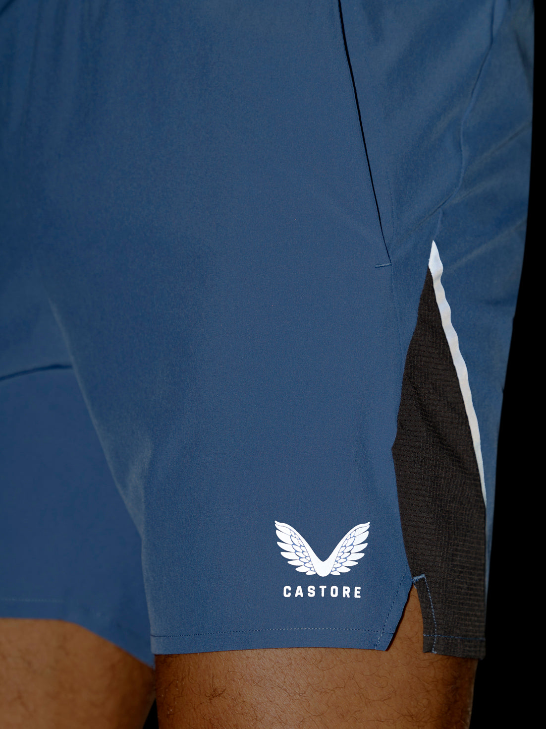 Close-up of blue athletic shorts worn by a model, featuring a white Castore wing logo on the lower left leg and a black mesh side panel with a white stripe.