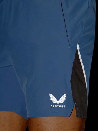 Close-up of blue athletic shorts worn by a model, featuring a white Castore wing logo on the lower left leg and a black mesh side panel with a white stripe.