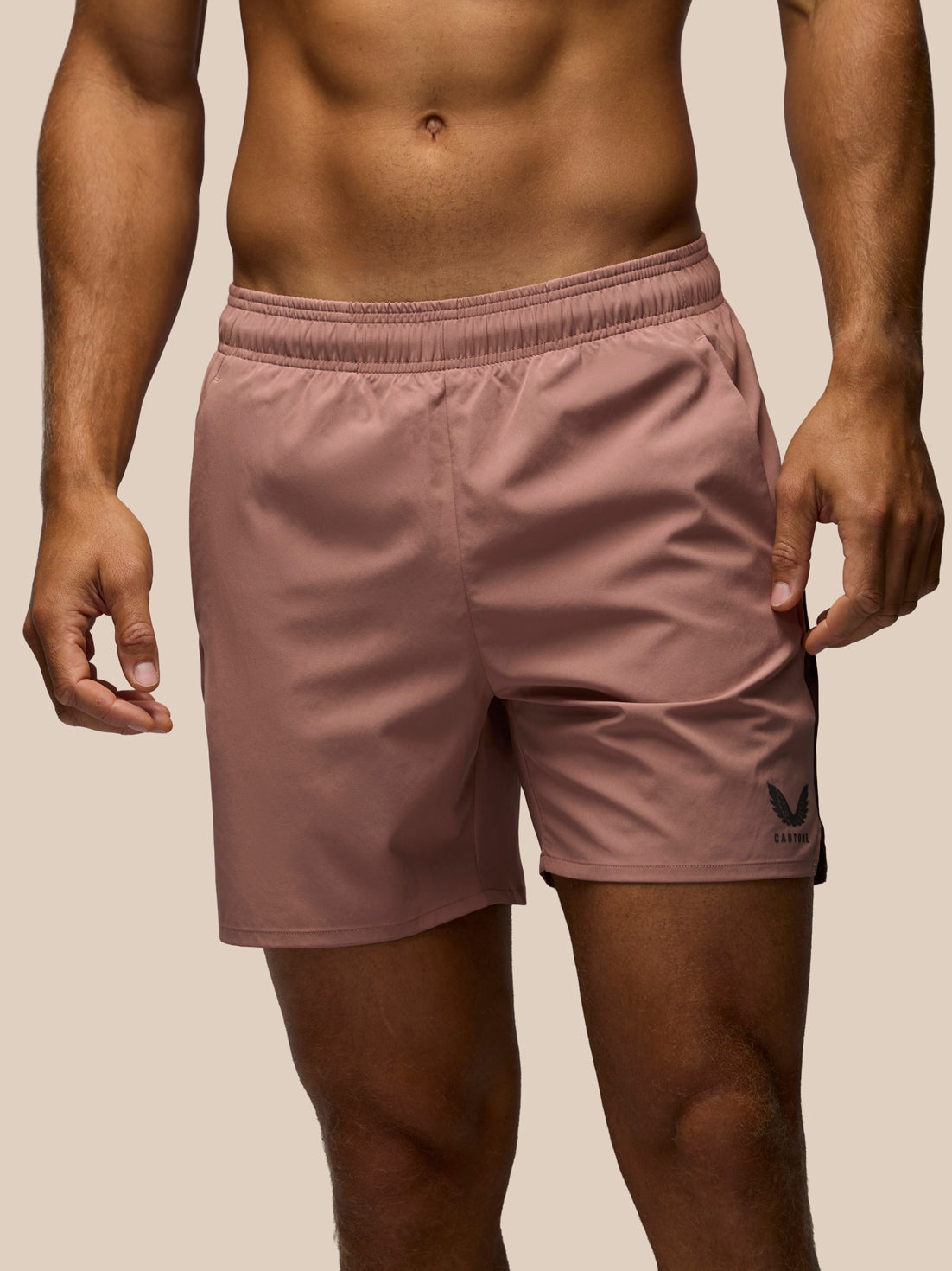 A close-up of a man wearing dusty rose athletic shorts with a black logo on the lower left leg, set against a solid beige background.