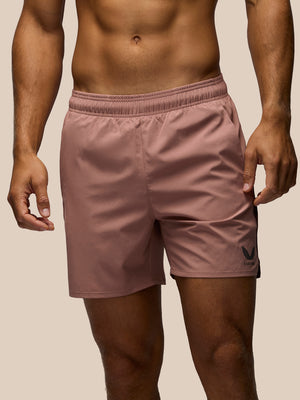 A close-up of a man wearing dusty rose athletic shorts with a black logo on the lower left leg, set against a solid beige background.