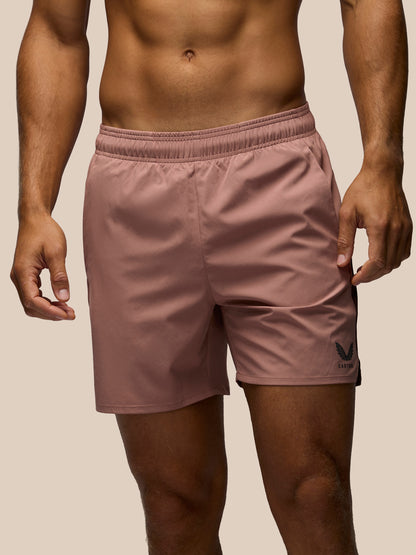 A close-up of a man wearing dusty rose athletic shorts with a black logo on the lower left leg, set against a solid beige background.