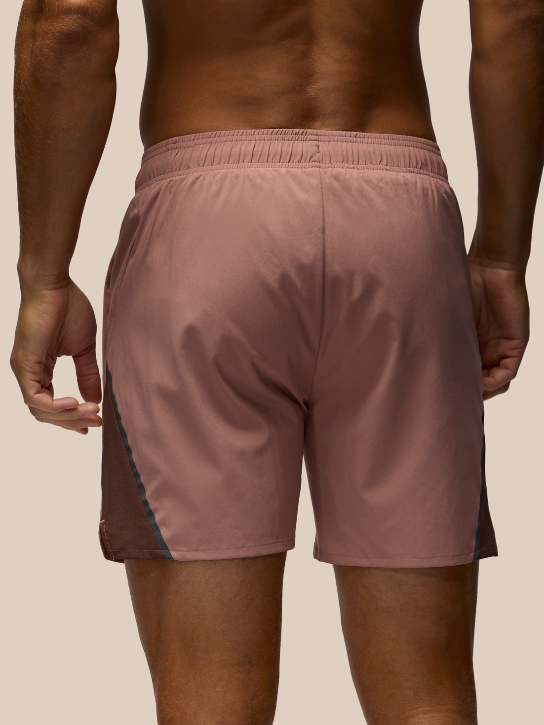 Rear view of a man wearing dusty rose athletic shorts with dark side panels and a thin teal stripe, against a light beige background.