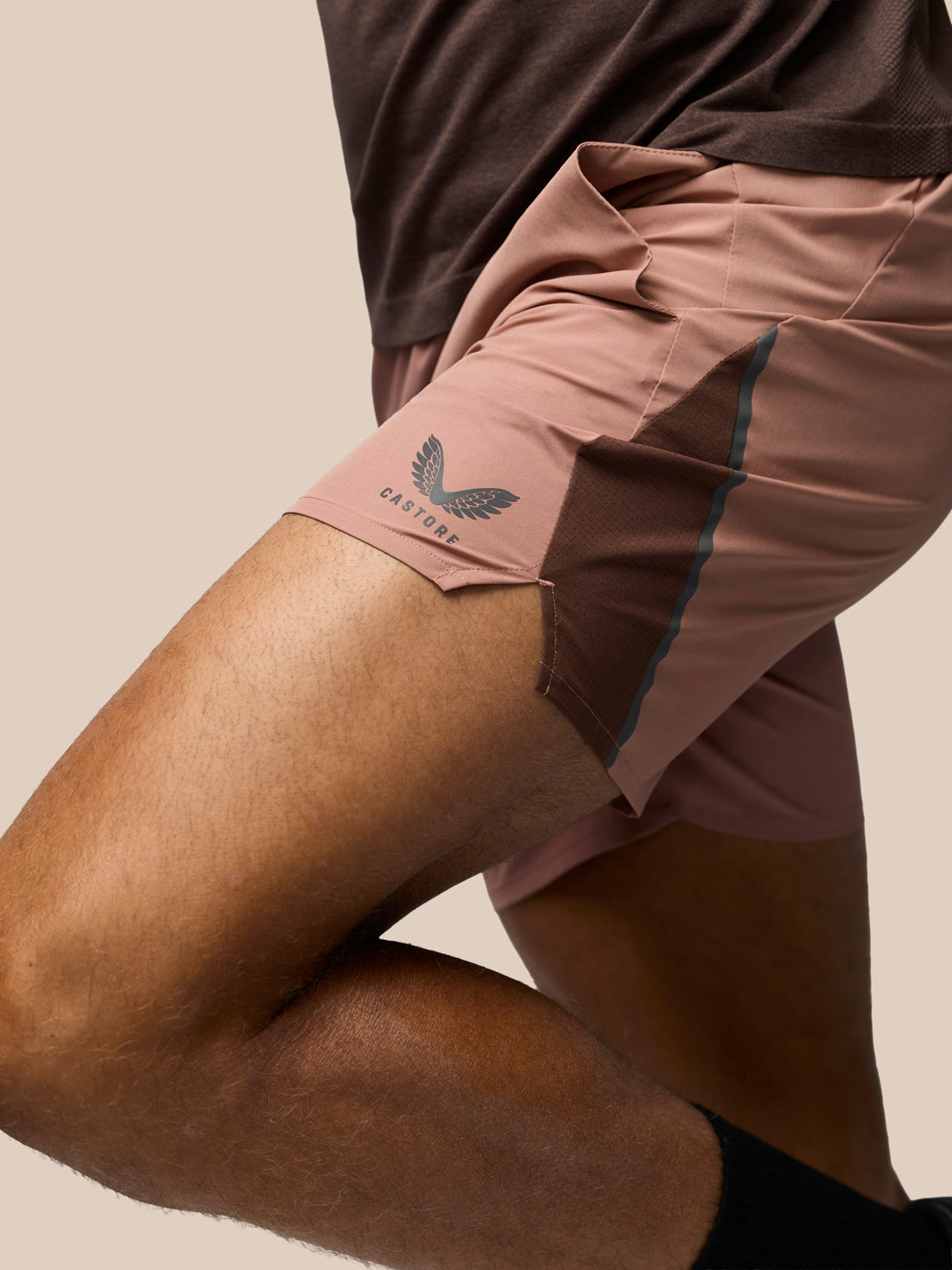 Close-up of a runner's legs wearing dusty rose and brown athletic shorts with a visible wing logo and the brand name 'CASTORE' on the side, against a plain beige background.
