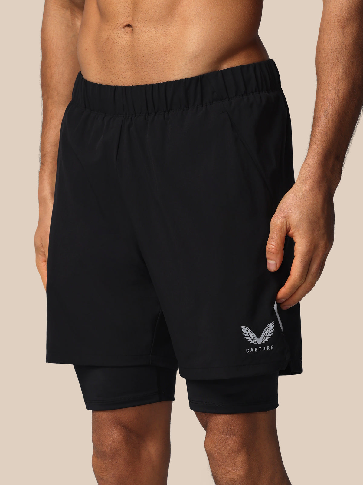 Black athletic shorts with a double-layered design, featuring a white wing logo and the brand name 'CASTORE' on the lower left leg, shown on a model against a solid beige background.