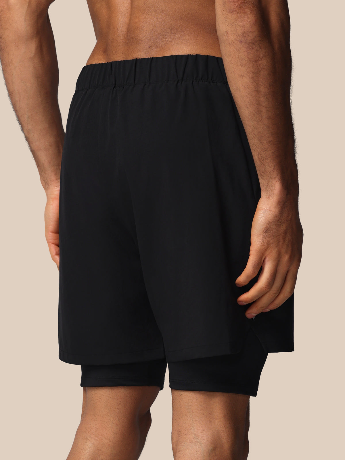 Black athletic shorts with an elastic waistband and a built-in second layer underneath, shown on a model from the back against a solid beige background.