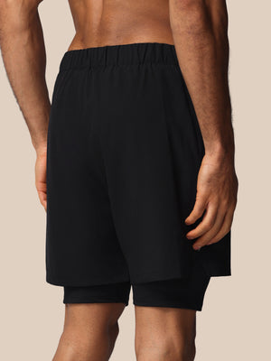 Black athletic shorts with an elastic waistband and a built-in second layer underneath, shown on a model from the back against a solid beige background.