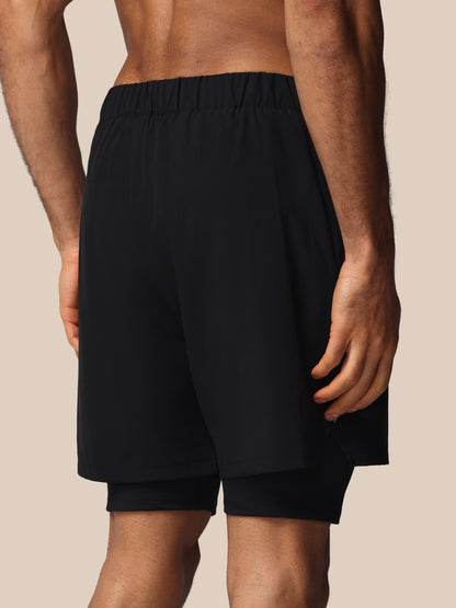 Black athletic shorts with an elastic waistband and a built-in second layer underneath, shown on a model from the back against a solid beige background.