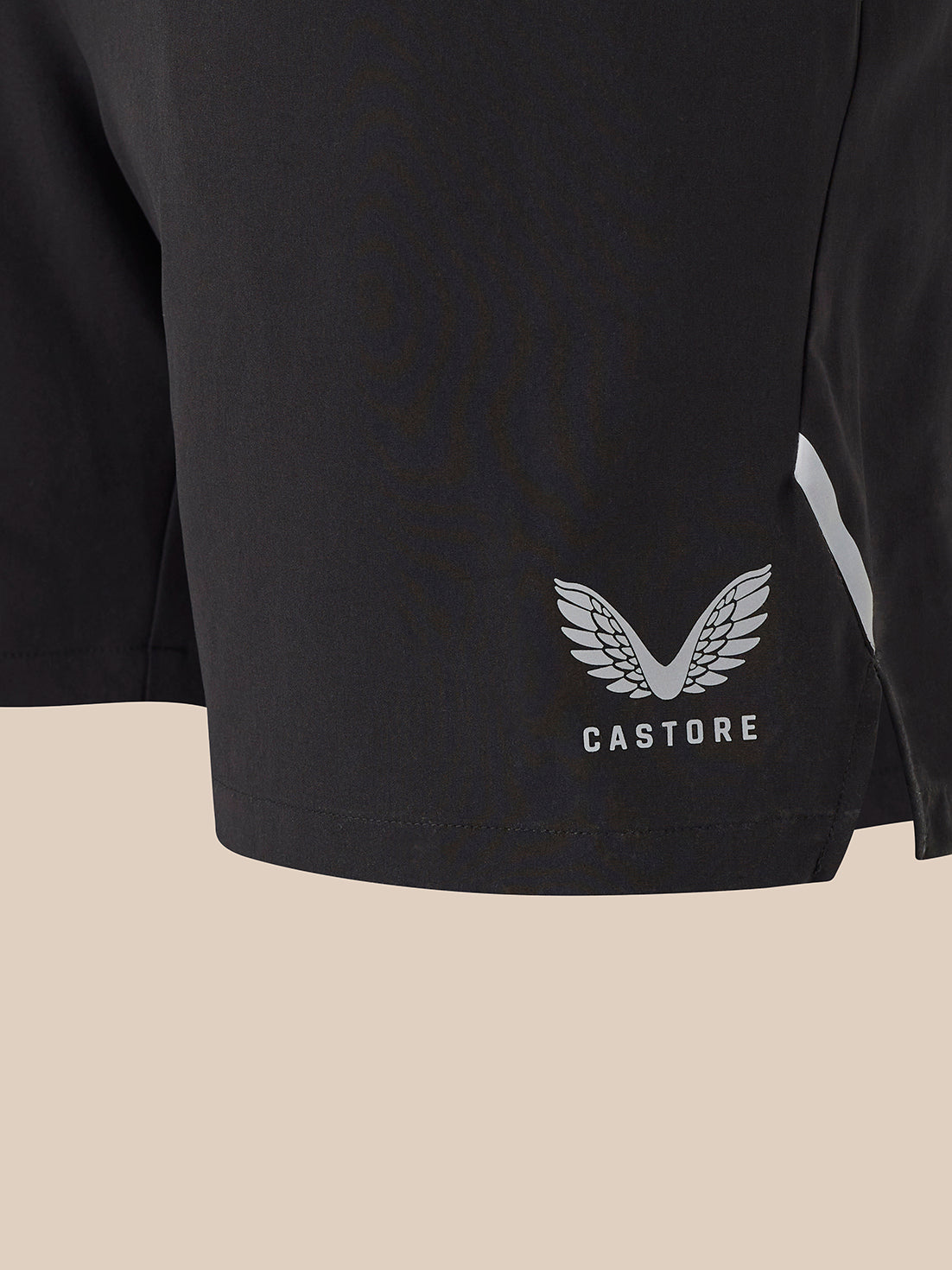 Close-up of a black athletic shorts leg featuring a white Castore wing logo and the brand name 'CASTORE' printed below it, set against a solid light beige background.