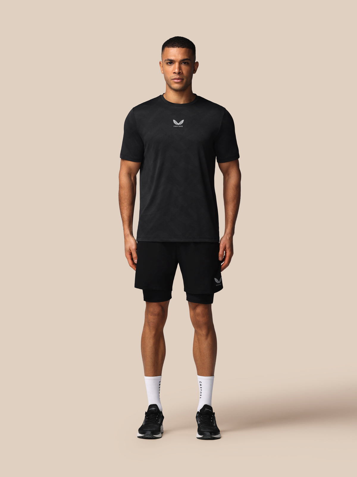 A man modeling a black short-sleeve athletic t-shirt and matching black shorts against a solid beige background.