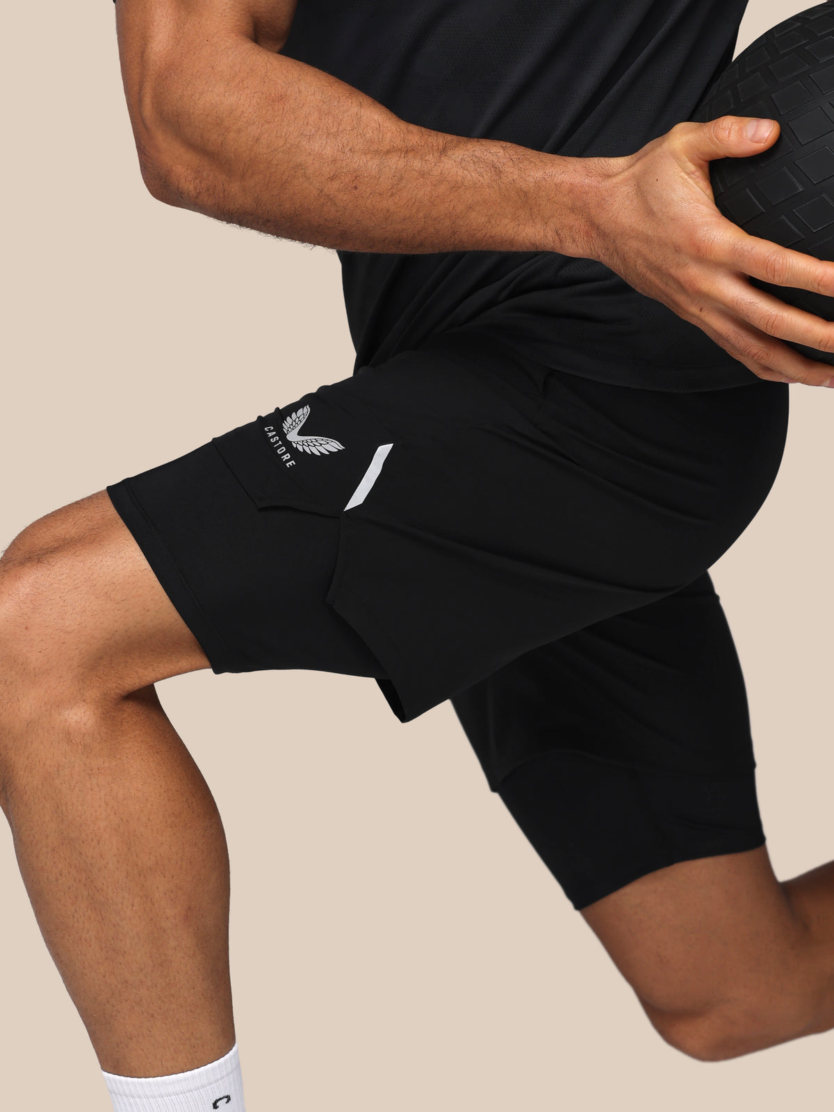 A close-up of a person wearing black athletic shorts with a white logo on the left leg, holding a black ball against a plain beige background.