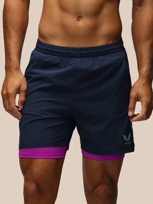A close-up of a man wearing navy blue athletic shorts with a purple inner layer peeking out at the hem, featuring a small white logo on the lower left leg.